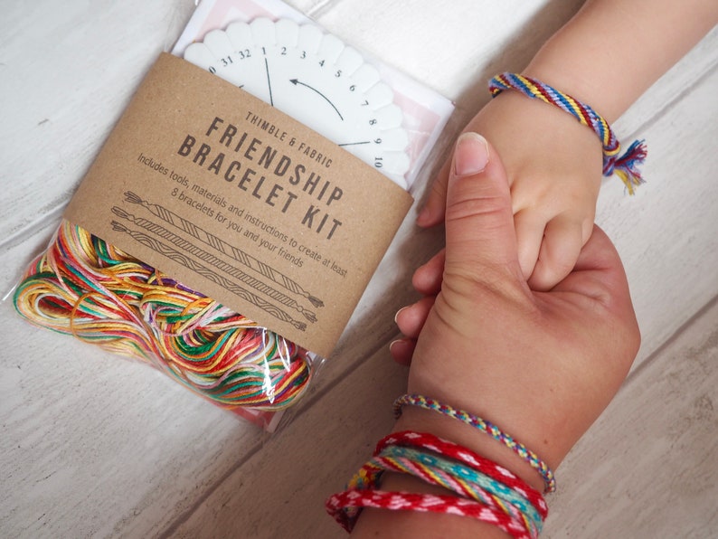Op de afbeelding: Een knutselset voor het maken van vriendschapsarmbanden. De set bevat kleurrijk borduurgaren, een plastic armbandweefraam en instructies. De set is verpakt in een bruine papieren zak met de tekst "Friendship Bracelet Kit" erop gedrukt.