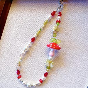 Puede incluir: Un collar caprichoso con un colgante de hongo rojo con lunares blancos, acentuado por hojas verdes y delicadas cuentas florales. El collar incorpora perlas blancas, cuentas rojas en forma de lágrima y varias cuentas verdes y transparentes, creando una estética de cuento de hadas.
