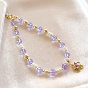 May include: A beaded phone charm featuring a repeating pattern of translucent light purple faceted beads, cream-coloured round beads, and small white ridged spacers. A gold-toned metal charm shaped like a cluster of flowers with a green leaf hangs from one end.