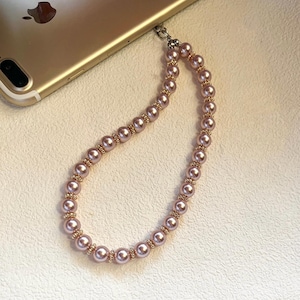 May include: A pink pearl phone strap with gold-colored accents. The strap is made of small, round, pink pearls strung together with gold-colored spacers. The strap is attached to a gold-colored phone.