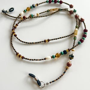 May include: A beaded eyeglass chain featuring a mix of small black seed beads interspersed with larger accent beads in shades of burgundy, emerald green, amber, white, and metallic gold, finished with black rubber loops and silver-toned metal connectors.