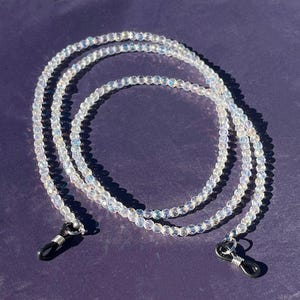 Peut inclure: Une chaîne de lunettes perlée transparente avec des boucles en métal argenté. La chaîne est composée de petites perles irisées qui captent la lumière. La chaîne de lunettes est présentée sur un fond violet foncé.