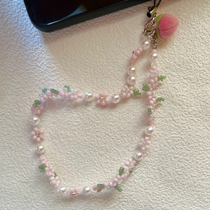 Puede incluir: Un charm de teléfono de cuentas rosas y verdes con un charm peludo de color melocotón adjunto. El charm está hecho de cuentas pequeñas y presenta un diseño floral.