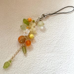 May include: A decorative charm featuring a cluster of beads and floral accents in shades of green, orange, yellow, and white. The charm includes a variety of bead shapes and sizes, with a silver-toned chain and a black cord loop for attachment.