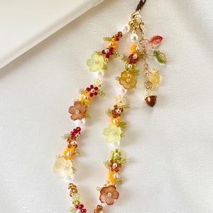 May include: A phone charm featuring a beaded loop with translucent flower-shaped beads in yellow, orange, and brown, interspersed with white faux pearls and small green beads. A gold-tone chain hangs from the top with leaf and acorn charms.