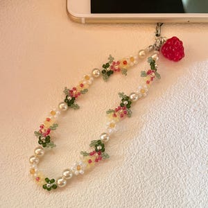May include: A white and pearl beaded phone charm with a floral design. The charm features small white, yellow, pink, and green beads arranged in the shape of flowers. A red plastic strawberry charm is attached to the end of the beaded chain.