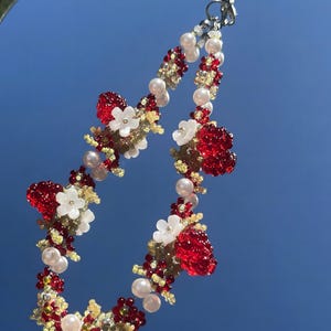 May include: A beaded phone charm featuring clusters of translucent red berry-like beads, white five-petaled flower beads, small pale yellow seed beads, and round white faux pearls. The charm is attached to a black cord with a silver metal lobster clasp.