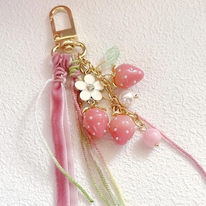 May include: A gold-coloured keychain featuring pink strawberry charms, a white flower, and a pearl. The keychain has pink and green braided ribbons and a pink velvet ribbon. The strawberries have white dot details.