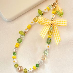 May include: A beaded phone charm featuring yellow and green glass beads, white daisy-shaped beads, and green leaf-shaped beads. The charm includes a yellow and white gingham ribbon bow and a small corn-shaped charm attached to a gold-tone clasp.