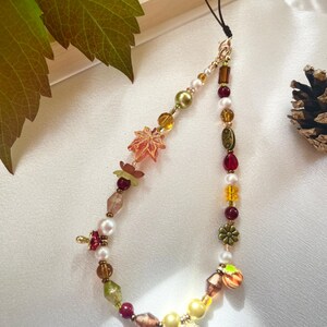 May include: A beaded phone charm featuring a variety of autumn-themed elements including a translucent orange maple leaf, a small acorn charm, and a flower-shaped bead. The strand is composed of white pearls, amber, red, and green beads.