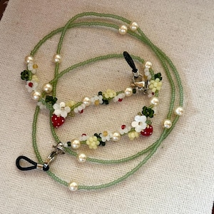 May include: A beaded spectacle chain featuring a long strand of small translucent green beads interspersed with cream-coloured faux pearls. The chain includes decorative segments of white and yellow flower beads and small red strawberry charms.