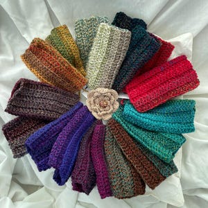 May include: A collection of colorful, handmade crochet headbands arranged in a circular pattern. The headbands display a variety of colors, including red, blue, green, and brown. A crocheted flower is in the center. The headbands are made of soft yarn.