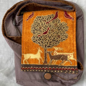 May include: A shoulder bag with a flap featuring a tree and deer design. The bag has a brown velvet strap and a button closure. The flap is orange with a tree and deer pattern, and the bag is a muted purple color.
