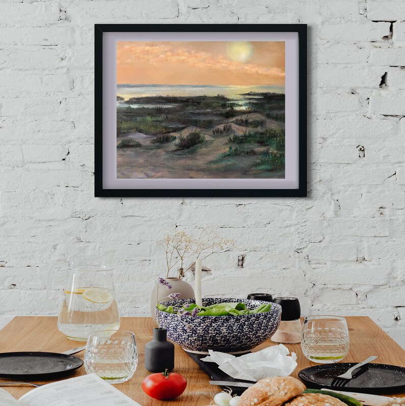 Pu&ograve; includere: Un dipinto incorniciato di un paesaggio con un tramonto su uno specchio d'acqua e dune di sabbia. Il dipinto &egrave; in una cornice nera ed &egrave; appeso a un muro di mattoni bianchi.