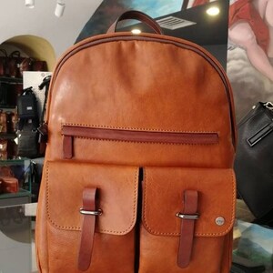 Könnte beinhalten: Ein brauner Lederrucksack mit zwei Fronttaschen und einer Reißverschlusstasche auf der oberen Klappe. Der Rucksack hat einen braunen Lederriemen und eine Schnalle an jeder Tasche.