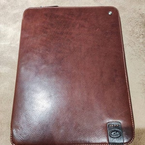 Può includere: Una custodia per laptop in pelle marrone con chiusura a zip e un piccolo logo in basso a destra.