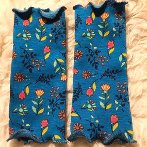 Könnte beinhalten: Zwei blaue Stoff-Armstulpen mit floralem Muster. Das Muster umfasst orange, rosa und gelbe Blumen mit grünen und schwarzen Blättern. Die Kanten sind mit einem Rüschenbesatz versehen.