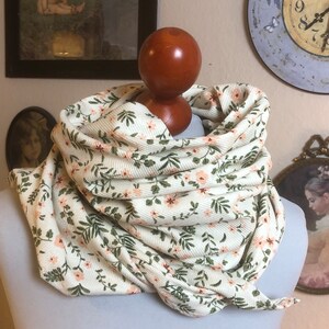 Könnte beinhalten: Ein weißer Schal mit einem Blumenmuster aus kleinen rosa Blüten und grünen Blättern. Der Schal ist gefaltet und um einen hölzernen Mannequin-Kopf gelegt.
