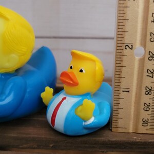 May include: Three rubber duck toys. The largest duck is yellow with a blue suit and a blonde wig. The middle duck is yellow with a blue suit and a blonde wig. The smallest duck is yellow with a red, white, and blue American flag design.