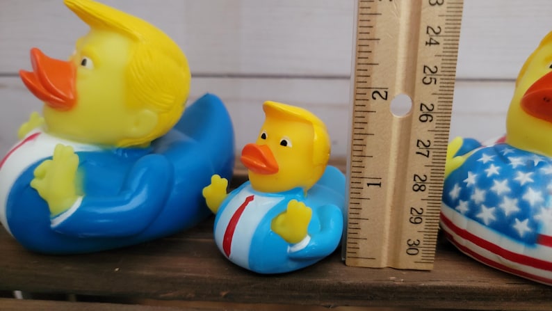 May include: Three rubber duck toys. The largest duck is yellow with a blue suit and a blonde wig. The middle duck is yellow with a blue suit and a blonde wig. The smallest duck is yellow with a red, white, and blue American flag design.