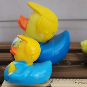 May include: Two yellow rubber ducks wearing blue suits. The larger duck has a yellow hairpiece and the smaller duck has a yellow hairpiece. The ducks are sitting on a wooden surface with a ruler.