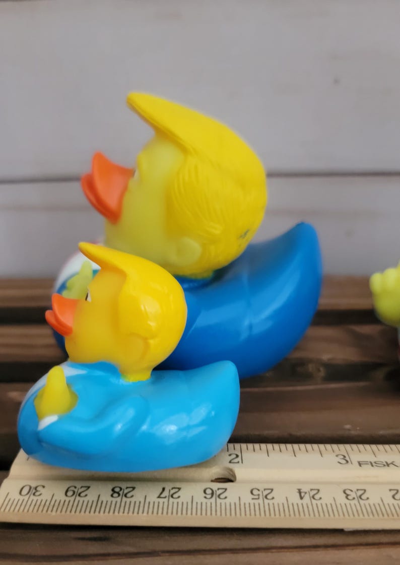 May include: Two yellow rubber ducks wearing blue suits. The larger duck has a yellow hairpiece and the smaller duck has a yellow hairpiece. The ducks are sitting on a wooden surface with a ruler.
