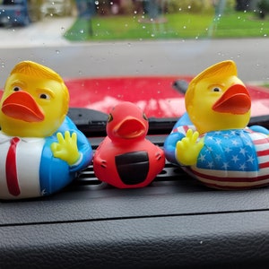 May include: Three rubber duck bath toys. One is yellow with a blue suit and red tie. Another is yellow with a red, white and blue American flag design. The third is red with a black beak.