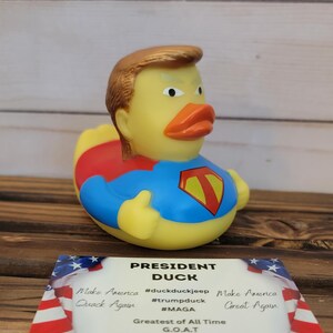 May include: A yellow rubber duck wearing a red and blue superhero costume, with a gold-coloured hairpiece and a serious expression. The duck is labelled "President Duck" with the text "Make America Great Again" and "Quack Again" on a card below it. The card also includes the hashtags #duckduckjeep, #trumpduck, and #MAGA.