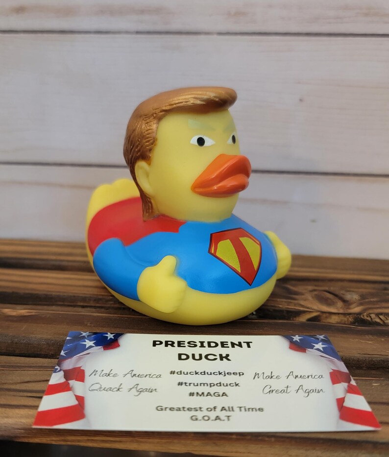 May include: A yellow rubber duck wearing a red and blue superhero costume, with a gold-coloured hairpiece and a serious expression. The duck is labelled "President Duck" with the text "Make America Great Again" and "Quack Again" on a card below it. The card also includes the hashtags #duckduckjeep, #trumpduck, and #MAGA.
