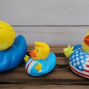 May include: Three rubber duck toys, one large and two smaller, with a yellow rubber duck head and a blue, red and white patterned body. The large duck has a blue body with a white collar and a red tie. The smaller ducks have a red, white and blue patterned body.