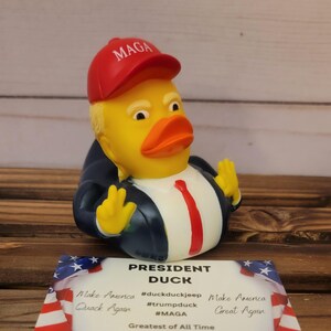 May include: A yellow rubber duck wearing a red baseball cap with the text "MAGA" on it. The duck is wearing a blue suit and a red tie. The duck is holding its hands up in a gesture of victory. The duck is sitting on a wooden surface. The text "President Duck" is written on a card in front of the duck. The card also includes the text "Make America Great Again" and "Greatest of All Time G.O.A.T."
