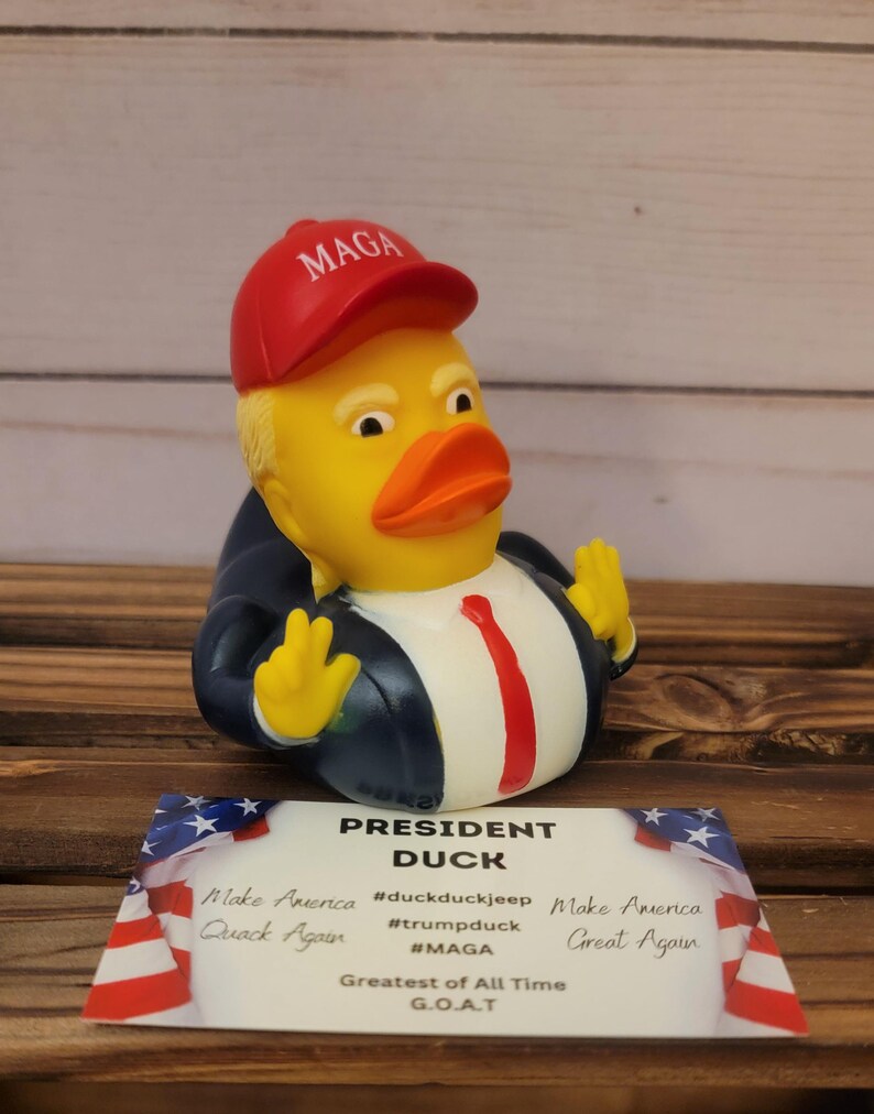May include: A yellow rubber duck wearing a red baseball cap with the text "MAGA" on it. The duck is wearing a blue suit and a red tie. The duck is holding its hands up in a gesture of victory. The duck is sitting on a wooden surface. The text "President Duck" is written on a card in front of the duck. The card also includes the text "Make America Great Again" and "Greatest of All Time G.O.A.T."