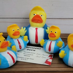 May include: A set of six yellow rubber duckies, each wearing a blue suit and red tie. The ducks have a yellow hairpiece resembling a comb-over. The largest duck has the text "President Duck" on a card in front of it. The card also includes the hashtags #duckluckjeep, #trumduck, and #MAGA.
