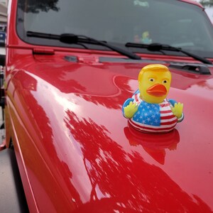 May include: A yellow rubber duck with a red, white and blue American flag design is perched on the bonnet of a red Jeep. The duck has a yellow beak and a yellow head with a blond hair design.