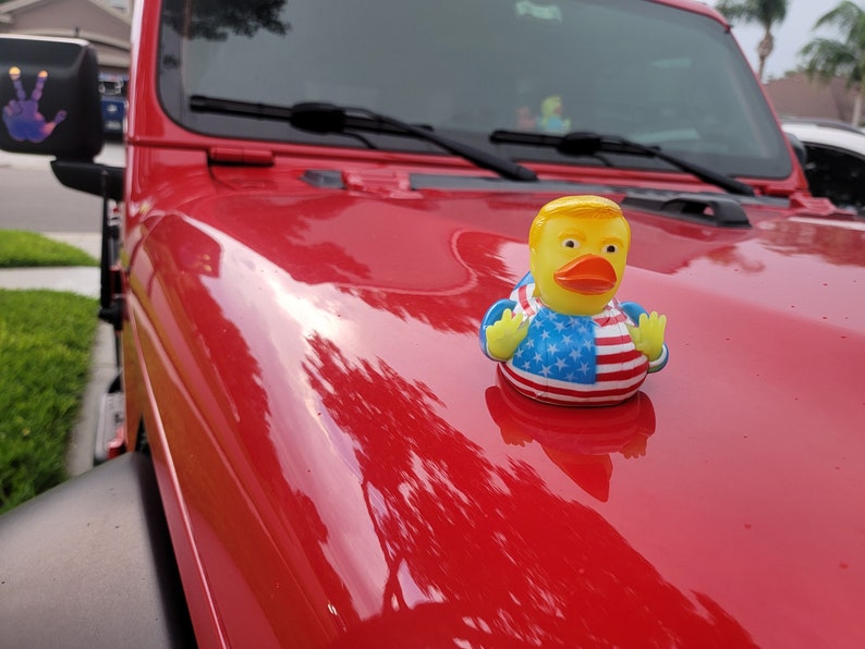 May include: A yellow rubber duck with a red, white and blue American flag design is perched on the bonnet of a red Jeep. The duck has a yellow beak and a yellow head with a blond hair design.
