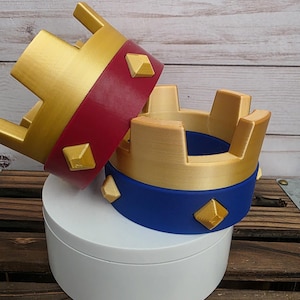 May include: Two decorative crowns, one red and gold, the other blue and gold, with gold diamond-shaped accents. The crowns are displayed on a white circular stand, set against a wooden background.