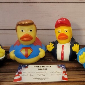May include: Four rubber duck toys, each depicting a different version of a political figure. The ducks are wearing suits and ties, with one wearing a red "MAGA" hat and another wearing a blue and red superhero costume. The ducks are arranged on a wooden surface with a sign that reads "President Duck, Greatest of All Time, GOAT."