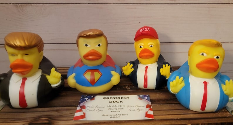 May include: Four rubber duck toys, each depicting a different version of a political figure. The ducks are wearing suits and ties, with one wearing a red "MAGA" hat and another wearing a blue and red superhero costume. The ducks are arranged on a wooden surface with a sign that reads "President Duck, Greatest of All Time, GOAT."