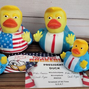 May include: Four rubber duck bath toys, each depicting a caricature of a president. The ducks are wearing blue suits and ties, and one is wearing a red, white and blue American flag design. The ducks are sitting on a wooden surface with a sign that reads "You've Been Ducked! President Duck".