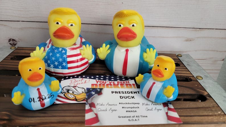May include: Four rubber duck bath toys, each depicting a caricature of a president. The ducks are wearing blue suits and ties, and one is wearing a red, white and blue American flag design. The ducks are sitting on a wooden surface with a sign that reads "You've Been Ducked! President Duck".