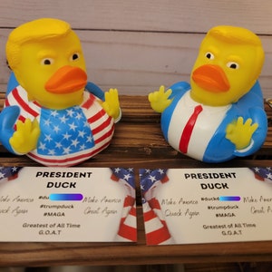 May include: Two yellow rubber duck toys wearing blue suits and red ties. One duck is wearing a red, white and blue American flag design. Both ducks have a card in front of them that says "President Duck" with the text "Make America Quack Again" and "Greatest of All Time G.O.A.T."
