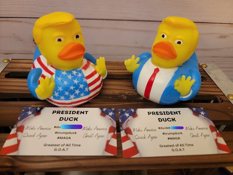 May include: Two yellow rubber duck toys wearing blue suits and red ties. One duck is wearing a red, white and blue American flag design. Both ducks have a card in front of them that says "President Duck" with the text "Make America Quack Again" and "Greatest of All Time G.O.A.T."