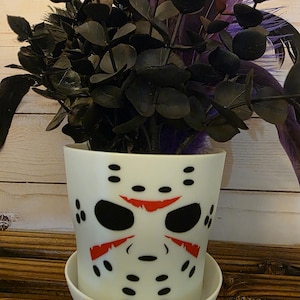 May include: A white ceramic planter with a black and red hockey mask design, filled with black faux eucalyptus and purple feathers. The planter sits on a matching white saucer, creating a spooky Halloween decoration.