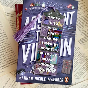 May include: A purple and black bookmark with a quote that reads "There is so much that can be fixed by honesty, if you're brave enough to use it." The bookmark is on top of a book titled "The Assassin's  Villain" by Hannah Nicole Maehrer.
