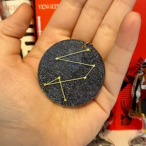 May include: A black glitter keychain with a gold constellation design. The constellation is a stylized version of the constellation Sagittarius.