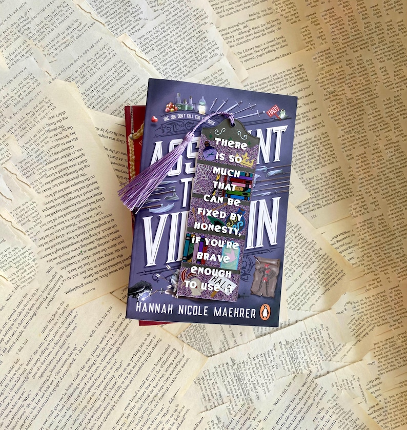 May include: A purple and black bookmark with a quote that reads "There is so much that can be fixed by honesty, if you're brave enough to use it." The bookmark is on top of a book titled "The Assassin's  Villain" by Hannah Nicole Maehrer.