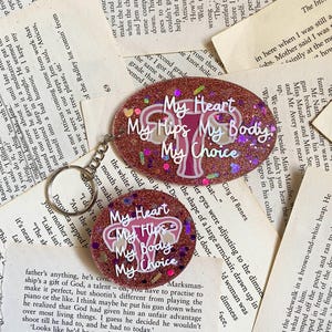 May include: Two round, pink and glitter resin keychains with a pink and white anatomical illustration of a uterus and the text "My Heart My Hips My Body My Choice".