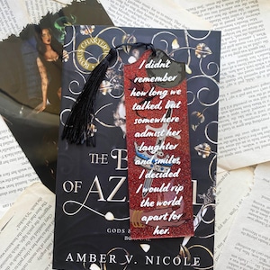 May include: A bookmark with a red glitter background and the text "I didn't remember how long we talked, but somewhere admist her..." is placed on a book. A black tassel hangs from the top. The book is titled "The Ballad of Azrael".