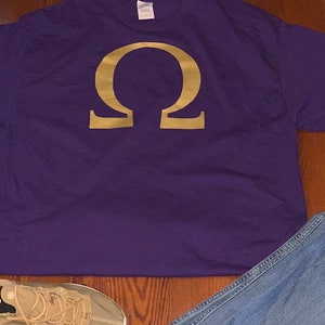 Puede incluir: Una camiseta morada con una letra griega omega dorada impresa en la parte delantera.