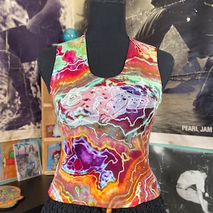 May include: A sleeveless tank top with a tie-dye pattern in shades of red, orange, yellow, purple, and blue. The tank top features a white embroidered lizard design. The tank top is on a black mannequin.